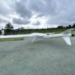 Side profile view of white long endurance hybrid VTOL fixed wing drone parked on outdoor test airfield ground, long range UAV for aerial mapping, land survey and infrastructure inspection