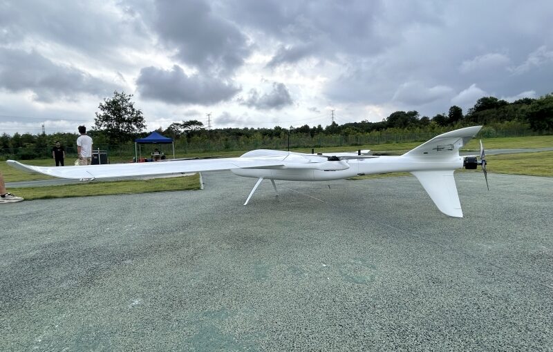 Side profile view of white long endurance hybrid VTOL fixed wing drone parked on outdoor test airfield ground, long range UAV for aerial mapping, land survey and infrastructure inspection