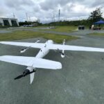 White hybrid VTOL fixed wing drone parked on the ground at outdoor test airfield, twin motor vertical takeoff landing UAV for aerial survey mapping and inspection missions
